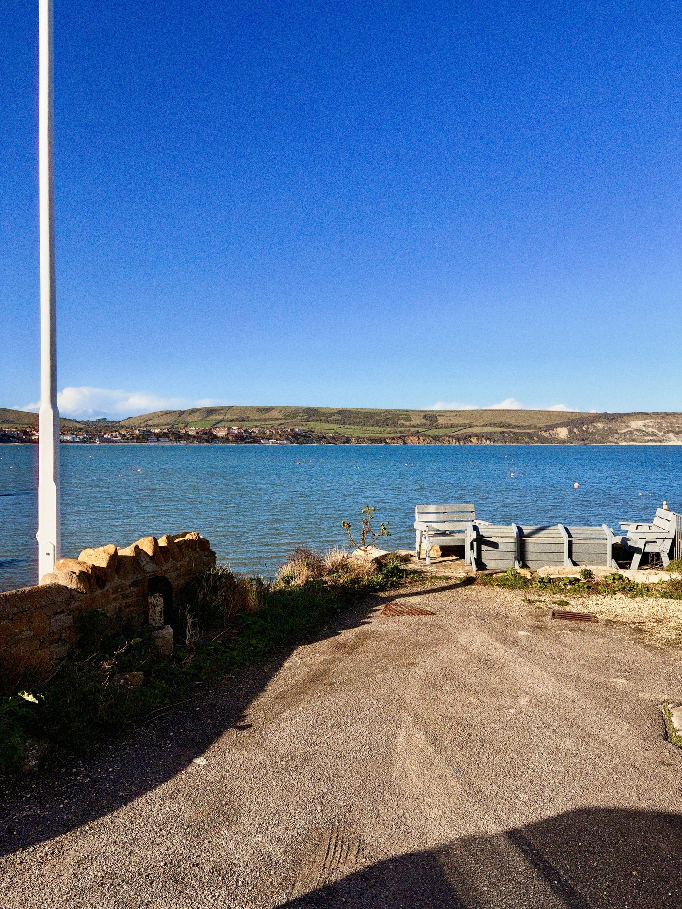A sunny bay with green hills and a seaside town across the water, and benches by a stone wall in the foreground