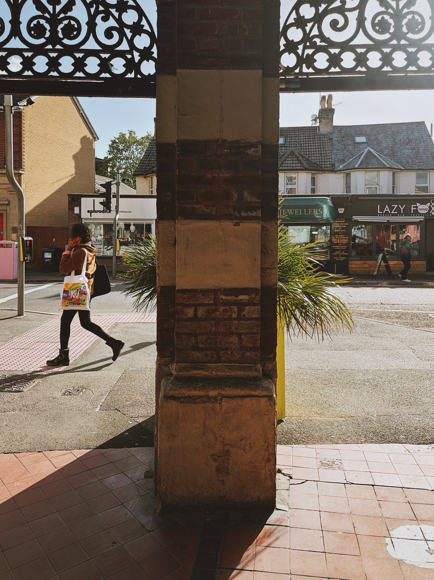 A brick pillar under an ornate iron canopy, with a woman walking past shops in the sunshine